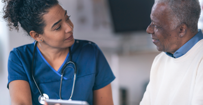 A female healthcare professional in a navy tunic speaking to an older man in a white jumper and shirt. 