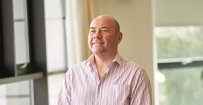 A photo of a tall white smiling man in a striped shirt and black jeans with his hands crossed in front of him. 