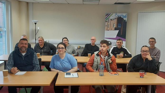 A group of ten people, diverse in age and gender, seated in a classroom-style setting and smiling towards the camera.